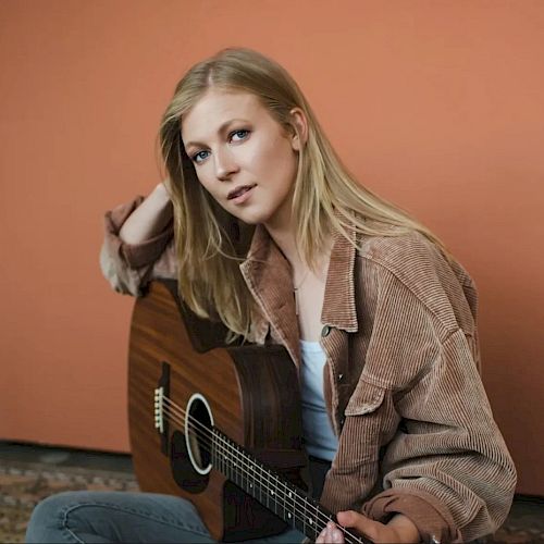 A person with long blonde hair is sitting against a peach-colored wall, wearing a brown shirt and holding an acoustic guitar, looking at the camera.