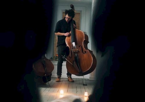 A person is shown playing a large string instrument, possibly a double bass, in a dimly lit room with candles on the floor, seen through a narrow gap.