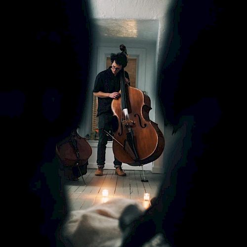 A person is shown playing a large string instrument, possibly a double bass, in a dimly lit room with candles on the floor, seen through a narrow gap.