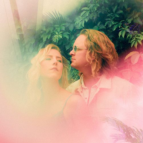 A man and woman stand close together, with vibrant, dreamy colors and foliage in the background, giving the image an ethereal, softly lit vibe.