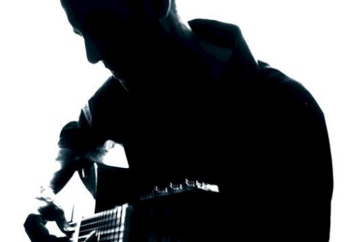 The image shows a silhouette of a person playing an acoustic guitar against a backlit background. The details of the person are not visible.