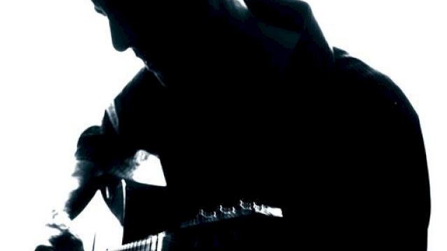 The image shows a silhouette of a person playing an acoustic guitar against a backlit background. The details of the person are not visible.