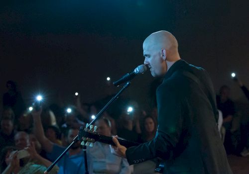 A bald musician plays a guitar and sings into a microphone on stage, with audience members holding up their phones, illuminating the dark venue.