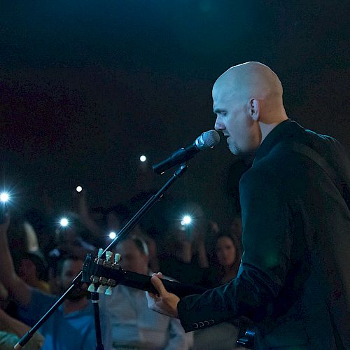 A bald musician plays a guitar and sings into a microphone on stage, with audience members holding up their phones, illuminating the dark venue.