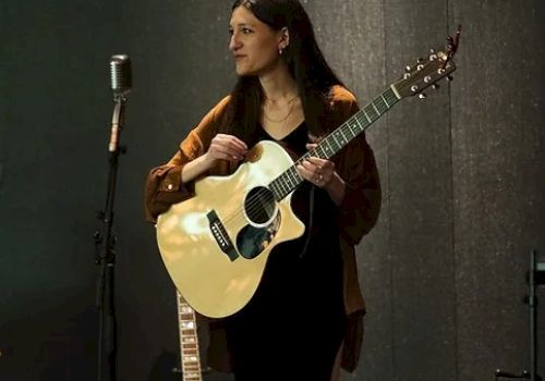 The image shows a woman holding an acoustic guitar, standing near a vintage microphone, with another guitar propped up against a wall in the background.