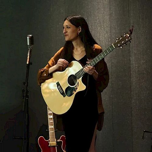 The image shows a woman holding an acoustic guitar, standing near a vintage microphone, with another guitar propped up against a wall in the background.