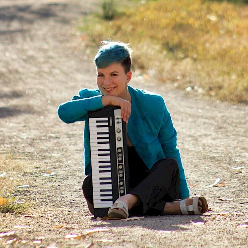 A person with blue hair is sitting on a path, smiling, and holding a small keyboard, leaning on it while dressed in a matching blue jacket and sandals.