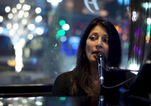 A woman is singing into a microphone, with colorful blurred lights visible in the background through a window.