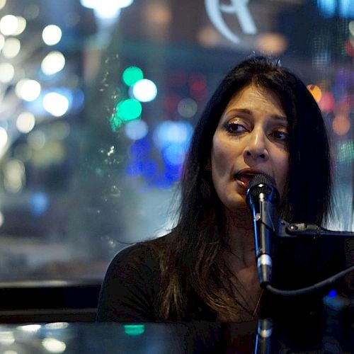 A woman is singing into a microphone, with colorful blurred lights visible in the background through a window.