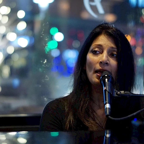 A woman is singing into a microphone, with colorful blurred lights visible in the background through a window.