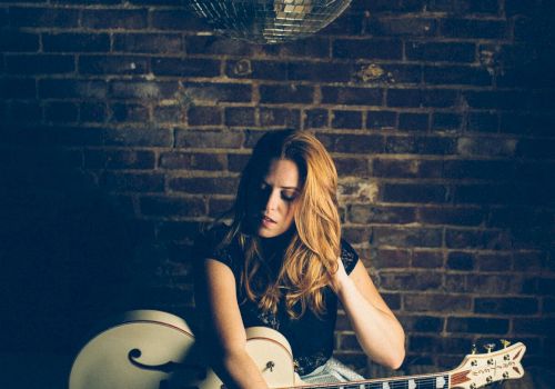 A person with long hair is seated, holding a guitar against a brick wall, with a disco ball hanging above.