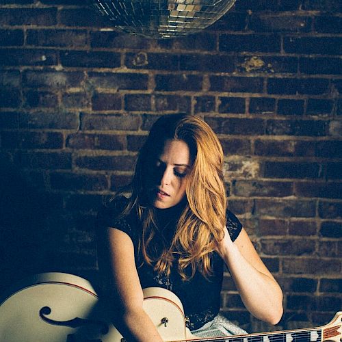 A person with long hair is seated, holding a guitar against a brick wall, with a disco ball hanging above.