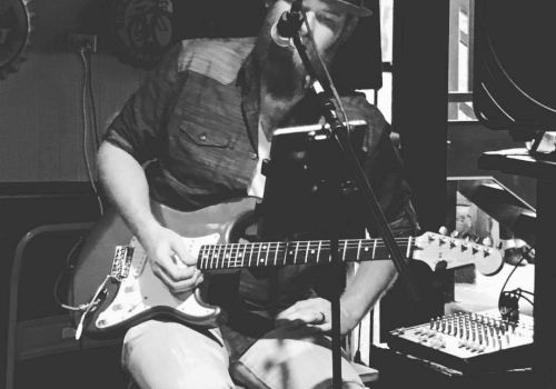 A bearded musician wearing a hat is sitting down, playing an electric guitar, and singing into a microphone in a dimly lit room.