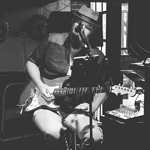 A bearded musician wearing a hat is sitting down, playing an electric guitar, and singing into a microphone in a dimly lit room.