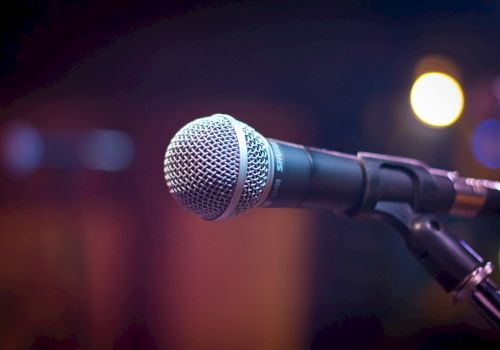 A close-up image of a microphone on a stand with blurred colorful lights in the background.