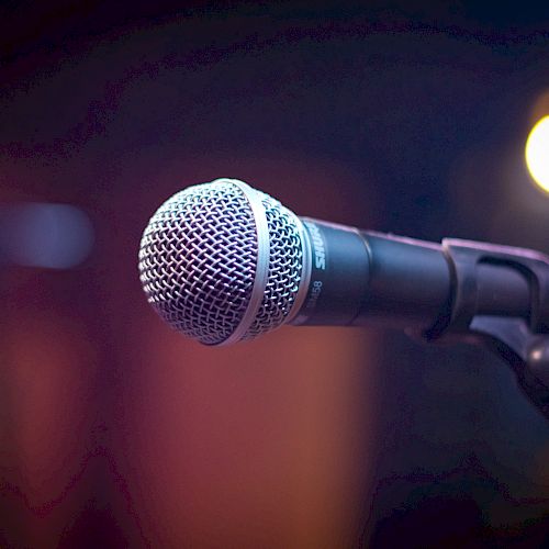 A close-up image of a microphone on a stand with blurred colorful lights in the background.