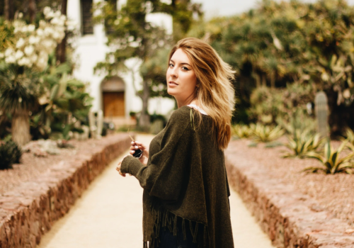 A woman stands on a path lined with trees and plants, looking back over her shoulder, with a white building in the background.