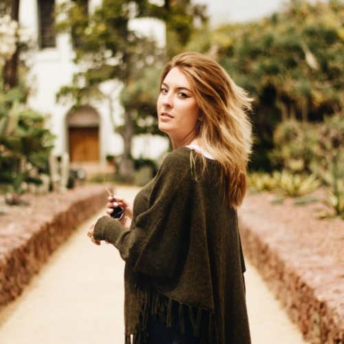 A woman stands on a path lined with trees and plants, looking back over her shoulder, with a white building in the background.