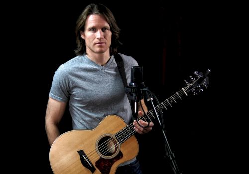 The image shows a person with long hair holding an acoustic guitar in front of a microphone, set against a black background.