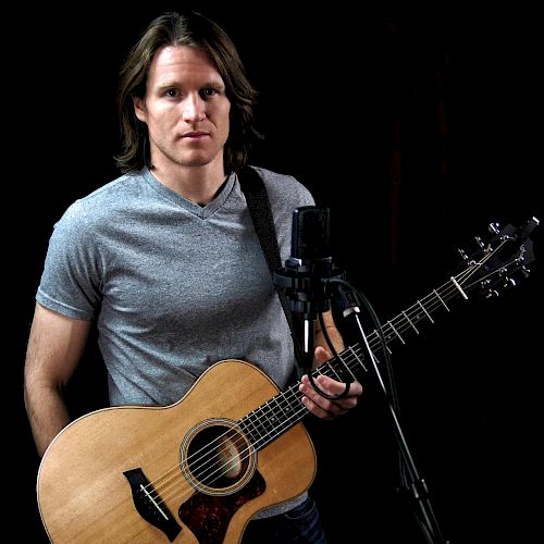 The image shows a person with long hair holding an acoustic guitar in front of a microphone, set against a black background.