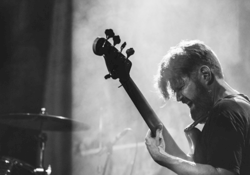 A person is passionately playing an electric bass guitar on stage with a drum set in the background, illuminated by stage lighting.