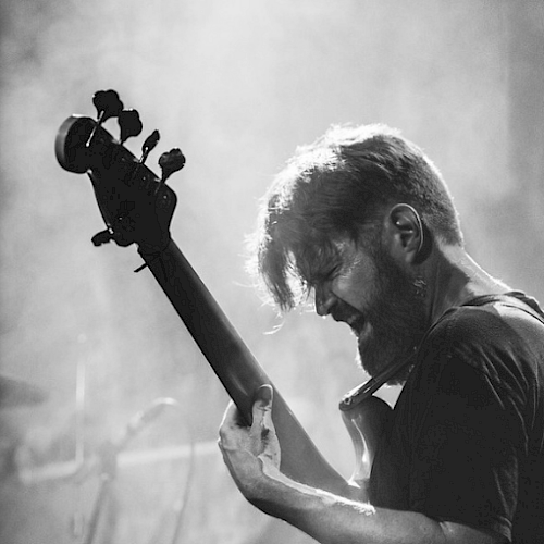 A person is passionately playing an electric bass guitar on stage with a drum set in the background, illuminated by stage lighting.