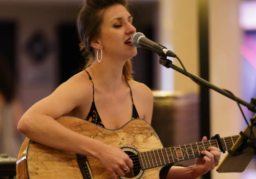 A woman is playing the guitar and singing into a microphone while seated in an indoor setting with a lamp in the background.