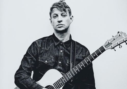 A black and white photo shows a person in a denim shirt holding an acoustic guitar against a plain background.