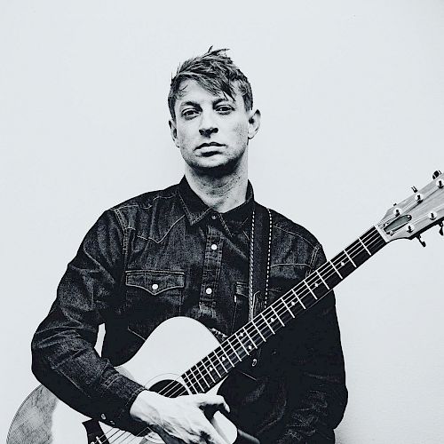 A black and white photo shows a person in a denim shirt holding an acoustic guitar against a plain background.