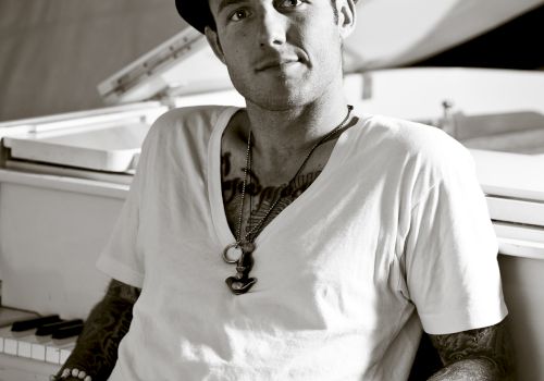 A man is sitting casually in front of a piano, wearing a hat and a white T-shirt.