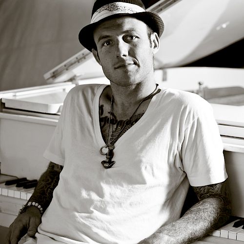 A man is sitting casually in front of a piano, wearing a hat and a white T-shirt.