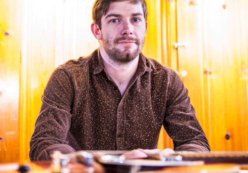 A man with a beard sits in front of a wooden wall, with several guitars blurred in the foreground.