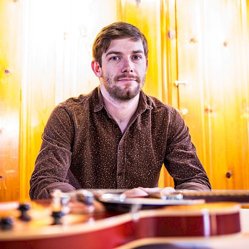 A man with a beard sits in front of a wooden wall, with several guitars blurred in the foreground.