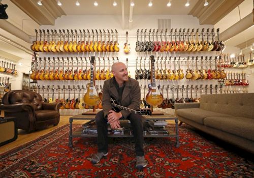 A person sitting in a room with numerous guitars displayed on wall racks, surrounded by a rug, a couch, and two chairs, smiling.