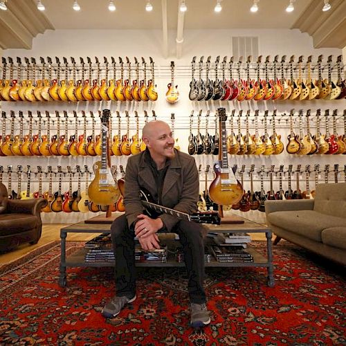 A person sitting in a room with numerous guitars displayed on wall racks, surrounded by a rug, a couch, and two chairs, smiling.