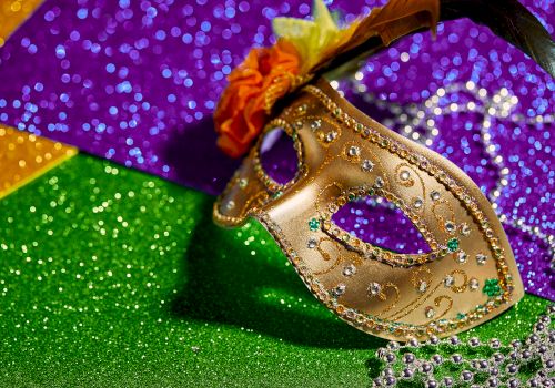 The image shows a decorated half-face mask, embellished with beads and flowers, set against a sparkling green, purple, and yellow backdrop, resembling Mardi Gras.