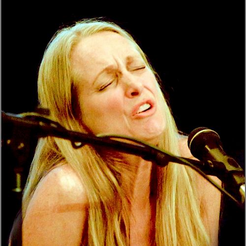 A person with long blonde hair appears to be passionately singing or performing, with a microphone in front of them, in a dark setting.