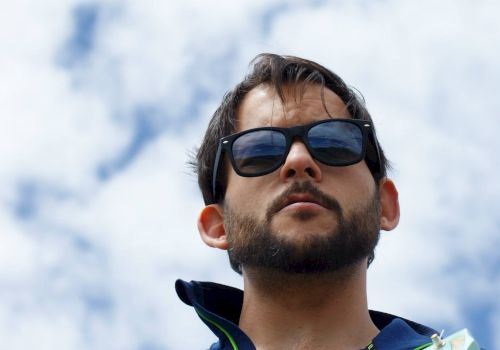 A man wearing sunglasses and a blue jacket is holding a guitar against a backdrop of a partly cloudy sky.