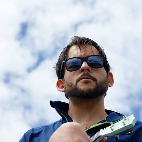 A man wearing sunglasses and a blue jacket is holding a guitar against a backdrop of a partly cloudy sky.