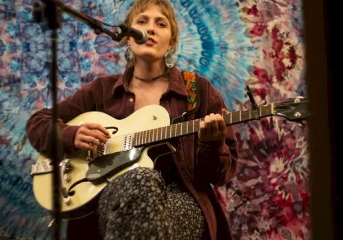 A person playing guitar and singing into a microphone, seated in front of a vibrant tie-dye tapestry backdrop, creating an artistic ambiance.