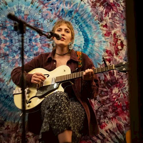 A person playing guitar and singing into a microphone, seated in front of a vibrant tie-dye tapestry backdrop, creating an artistic ambiance.