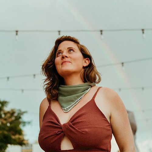 A person with a brown top and green scarf looks up, with string lights and a faint rainbow in the background under a cloudy sky.