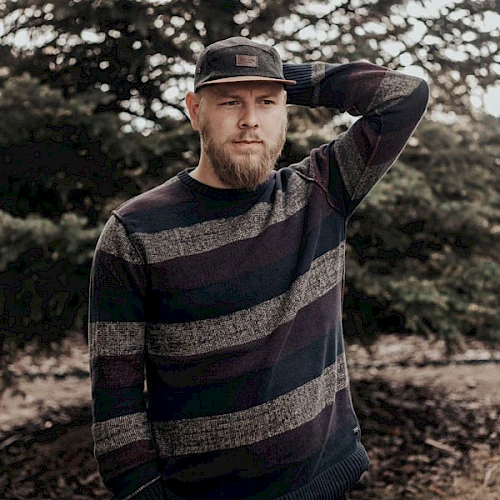 A person with a beard wearing a striped sweater and cap is standing outdoors near trees, with one hand on their head.