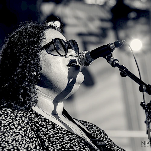 A person with curly hair and sunglasses sings into a microphone on stage, as seen in a black-and-white photograph.