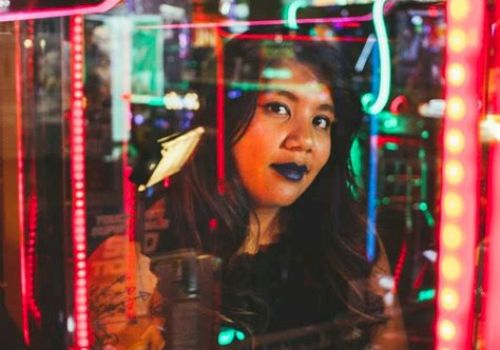 A woman is seen through colorful neon lights, reflecting an urban, vibrant atmosphere.