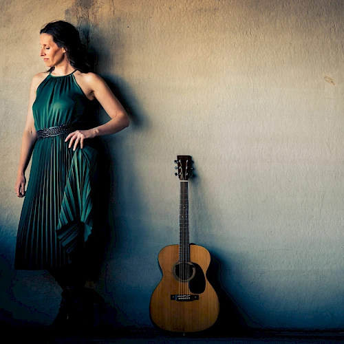 A woman in a green dress stands against a wall, with a guitar leaning beside her.
