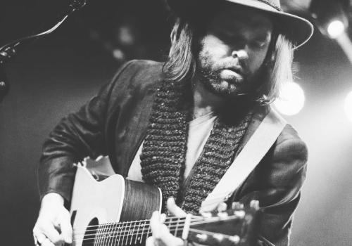 A person playing an acoustic guitar on stage, wearing a hat and scarf, with a microphone nearby, illuminated by stage lights.