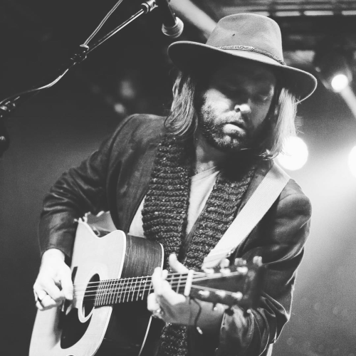 A person playing an acoustic guitar on stage, wearing a hat and scarf, with a microphone nearby, illuminated by stage lights.