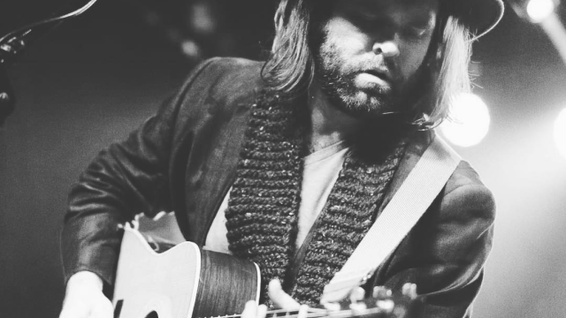 A person playing an acoustic guitar on stage, wearing a hat and scarf, with a microphone nearby, illuminated by stage lights.