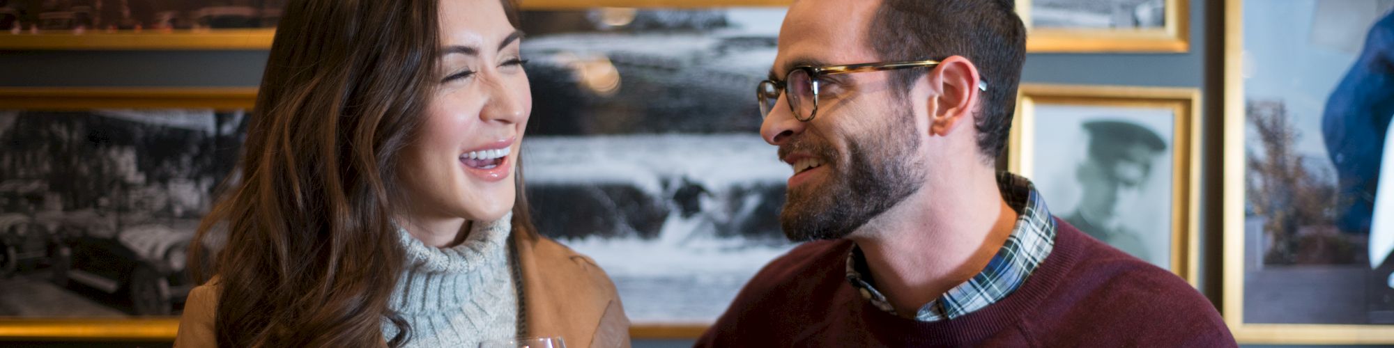 A woman and a man are chatting and laughing over drinks in a cozy setting, with framed photos on the wall in the background.